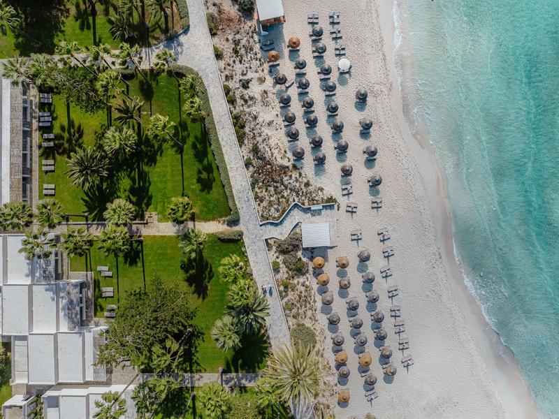 Drohnenansicht eines Strandes mit Sonnenschirmen und angrenzendem grünen Hotelgelände.