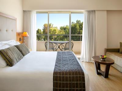 Helles Hotelzimmer mit großem Bett, Balkon und Blick auf grüne Bäume, Sofa und kleinem Holztisch.