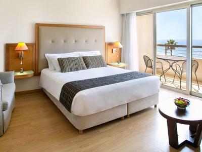 Helles Schlafzimmer mit Doppelbett, Holzboden, Balkon und Meerblick, zwei Nachttischen und Sessel.