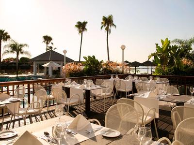 Terrazza soleggiata con tavoli apparecchiati, vista su piscina e palme sotto cielo limpido.