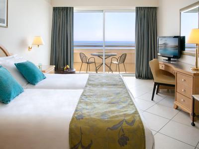 Habitación de hotel luminosa con cama doble, balcón con vista al mar y muebles de madera modernos.
