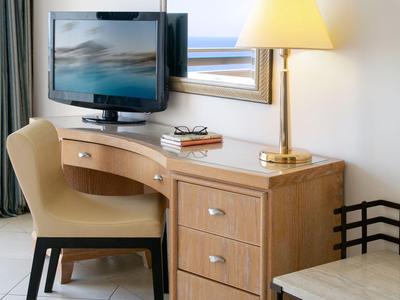 Esquina de madera clara con silla, TV, espejo y lámpara contra pared blanca en habitación de hotel.