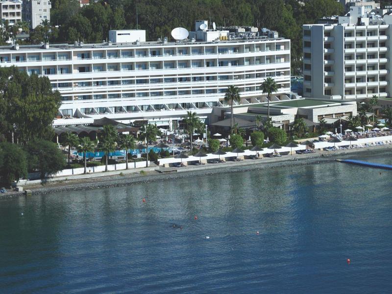 Großes Hotel am Wasser mit mehreren Pools und Liegestühlen, umgeben von grünen Bäumen.