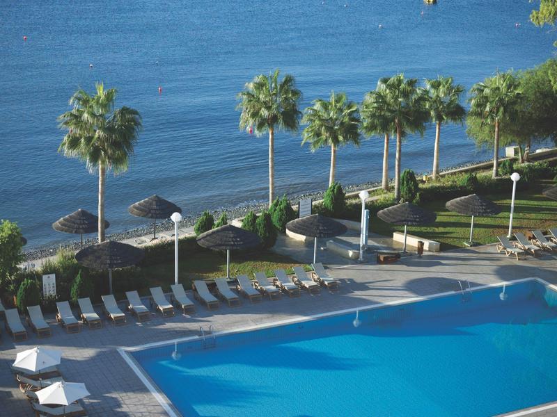 Großer Pool mit Liegestühlen, Palmen und Blick auf ruhiges, blaues Meer am Ufer.