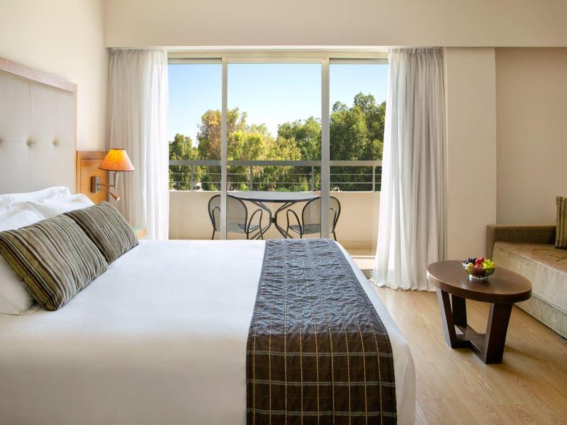 Helles Hotelzimmer mit großem Bett, Balkon und Blick auf grüne Bäume, Sofa und kleinem Holztisch.