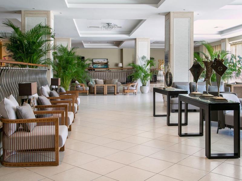 Großer, heller Lounge-Bereich mit weißen Wänden, Holzmöbeln, Kissen und vielen grünen Pflanzen.