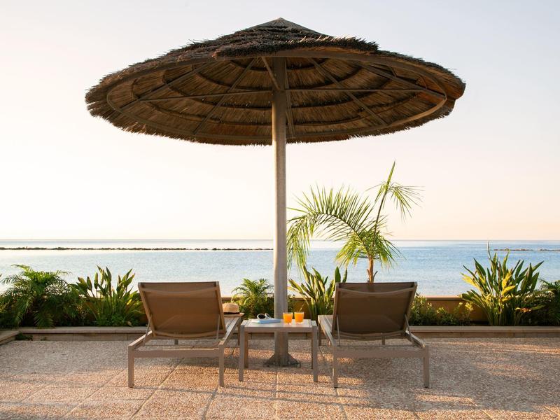 Zwei Liegestühle unter einem großen Strohmantelschirm mit Blick auf das Meer bei Sonnenuntergang.