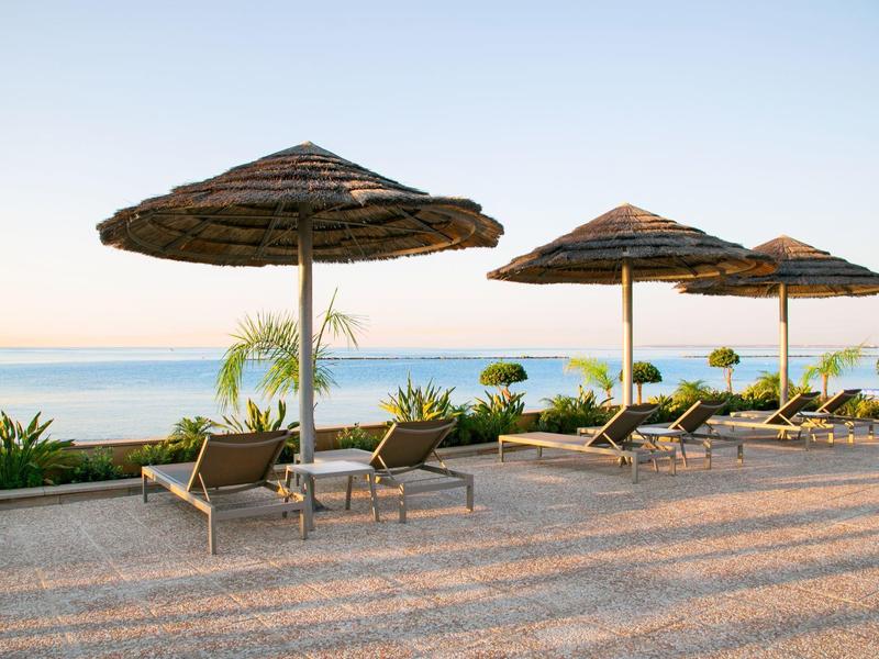 Strandliegen mit Strohschirmen neben ruhigem Meer und klar blauem Himmel bei Sonnenaufgang.