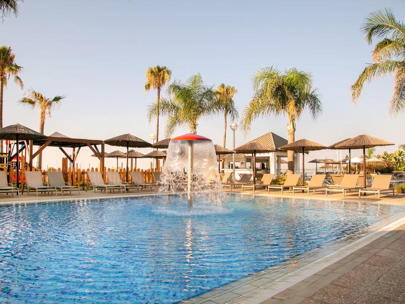 Großer Pool mit Springbrunnen, Palmen und Sonnenschirmen unter klarem Himmel.