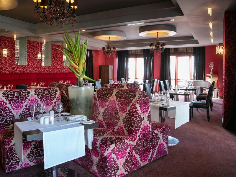 Elegantes Restaurant mit roten gemusterten Sesseln, weißen Tischen und großen Fenstern mit Vorhängen.