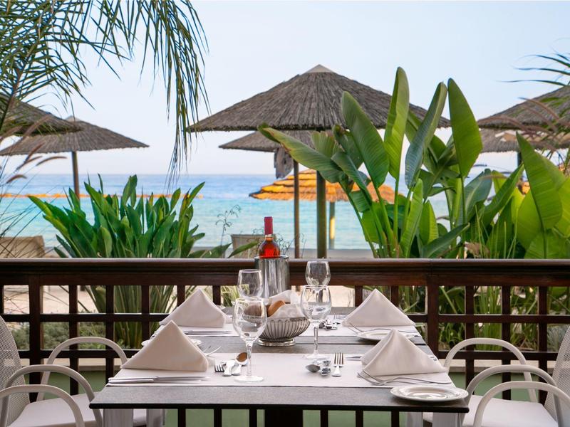 Restaurantterrasse mit eingedecktem Tisch, weißen Stühlen, grünen Pflanzen und Strand mit Sonnenschirmen.