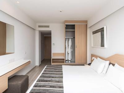 Helles Schlafzimmer mit Doppelbett, grauem Teppich, Holzschrank und großem Spiegel an weißer Wand.