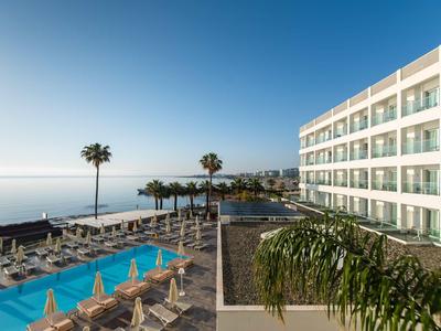 Modernes Hotel mit Außenpool, Liegestühlen und Blick auf das Meer bei klarem Himmel.