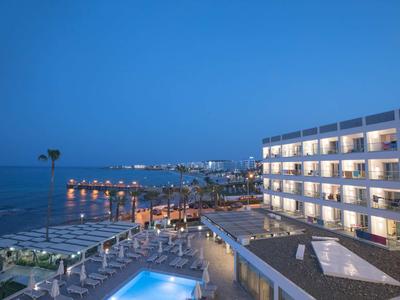 Nächtliche Ansicht eines beleuchteten Hotels am Meer mit Pool und Liegestühlen.