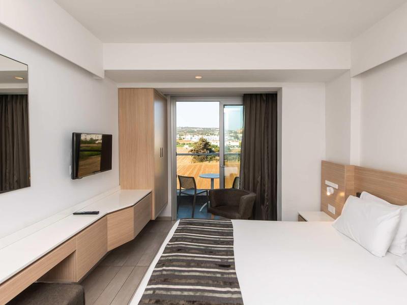 Modernes Hotelzimmer mit großem Bett, Schreibtisch, Flachbildfernseher und Balkon mit Sicht nach außen.