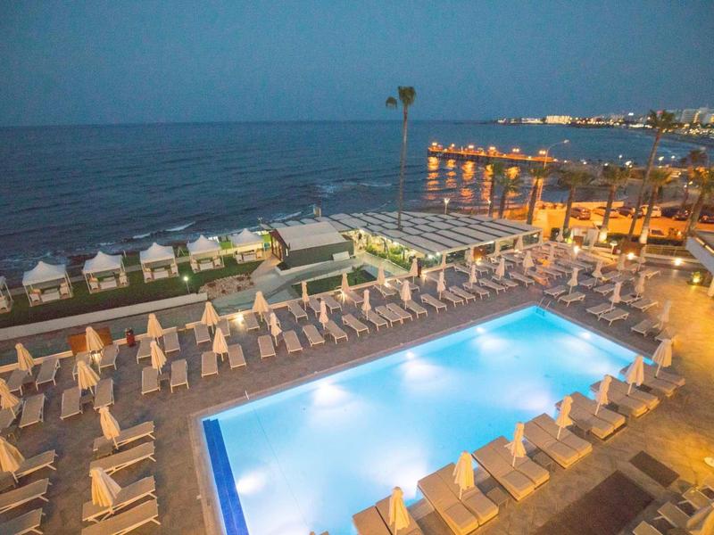 Beleuchteter Pool mit weißen Liegen und Sonnenschirmen am Meer bei Abenddämmerung.