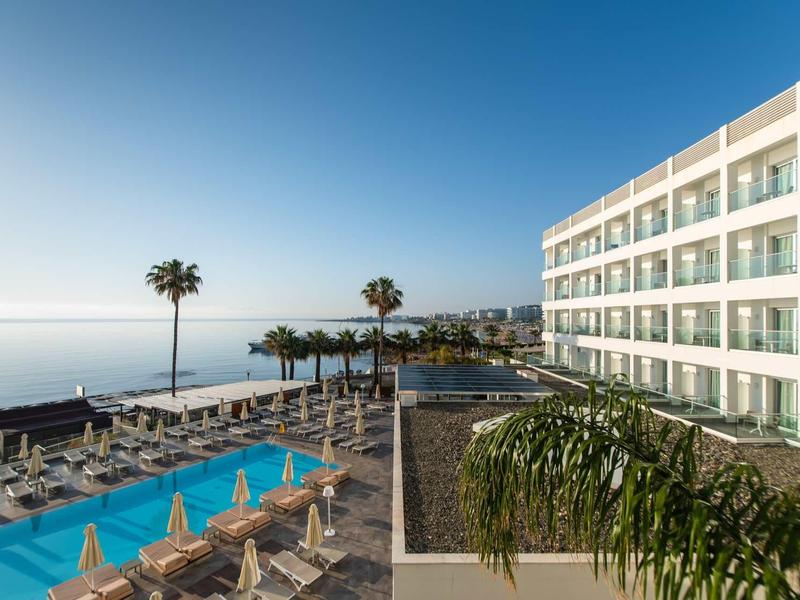 Moderner Hotelkomplex mit Pool, Liegestühlen, Palmen und Blick auf das ruhige, blaue Meer.