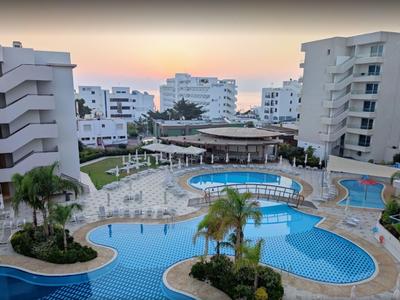 Hotelanlage mit mehreren Pools, Sonnenliegen und umliegenden mehrstöckigen Gebäuden bei Sonnenuntergang.