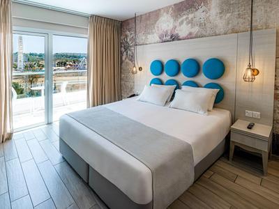 Helles Hotelzimmer mit Doppelbett, blauen Kopfkissen und Balkon mit Ausblick.