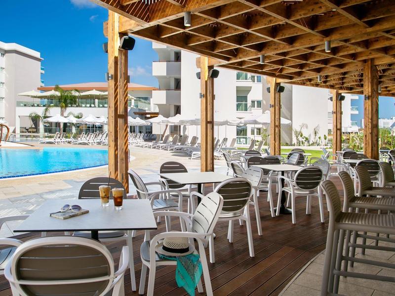 Überdachte Terrasse mit weißen Tischen, Stühlen und Blick auf Hotelpool und Gebäude bei klarem Himmel.