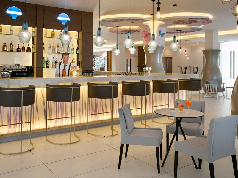 Moderner Barbereich mit hohem beleuchtetem Tresen, schwarzen Hockern und weißen Stühlen an Tischen.