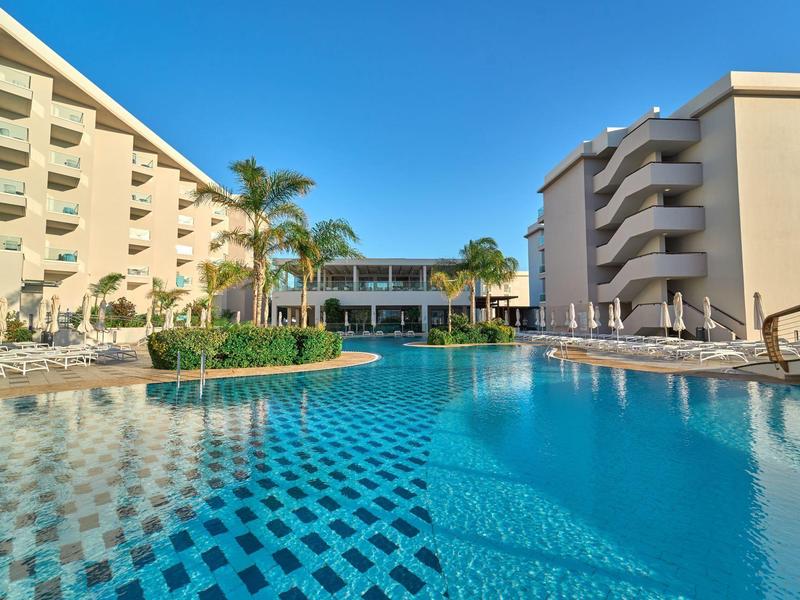 Großer Pool mit blauen Fliesen zwischen zwei modernen Hotelgebäuden unter klarem Himmel.