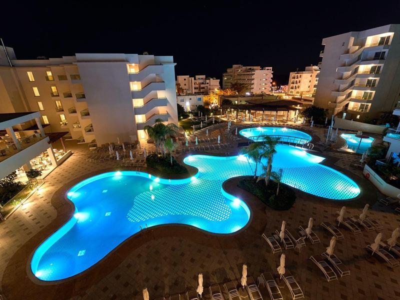 Beleuchteter Pool mit organischer Form, umgeben von Hotelgebäuden und Liegestühlen bei Nacht.