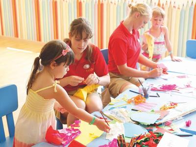 Kinder und Erzieherinnen basteln gemeinsam an einem Tisch mit buntem Papier und Scheren.