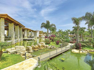 Luxuriöse Hotelanlage mit Teich, Fußgängerbrücke, Palmen und blühenden Pflanzen unter blauem Himmel.