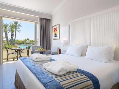 Helles Schlafzimmer mit großem Bett, weißen Wänden, blauer Bettdecke und Balkon mit Blick auf Palmen.