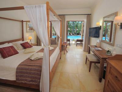 Helles Hotelzimmer mit Himmelbett, Sitzbereich und Blick auf einen Außenpool durch große Fenster.