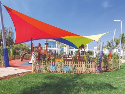 Bunter Sonnensegel-Schattenspender über einem eingezäunten Spielplatz mit Rutschen und Palmen.