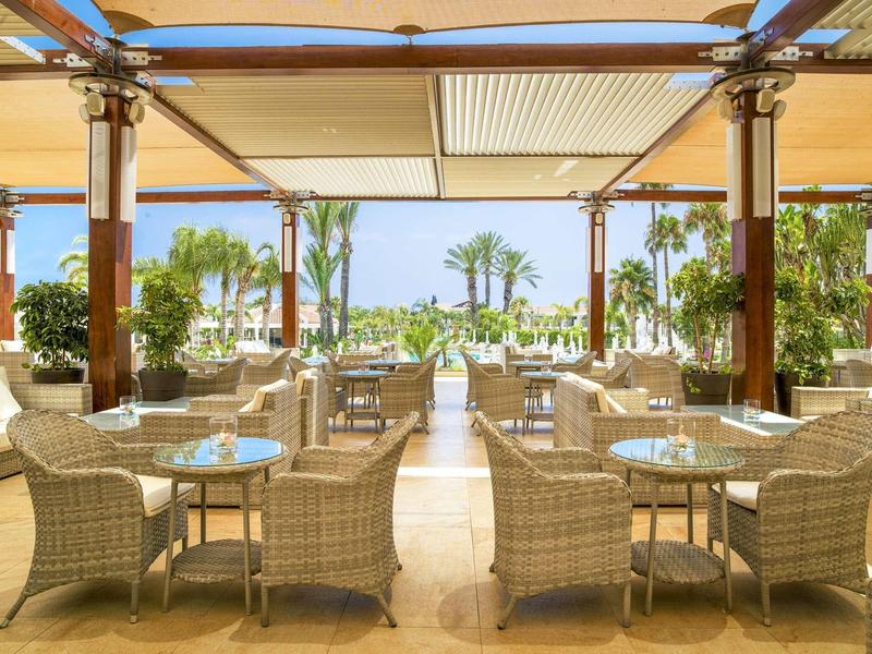 Hell eingerichtete Terrasse mit Korbsesseln und Glastischen, Blick auf Palmen und blauen Himmel.