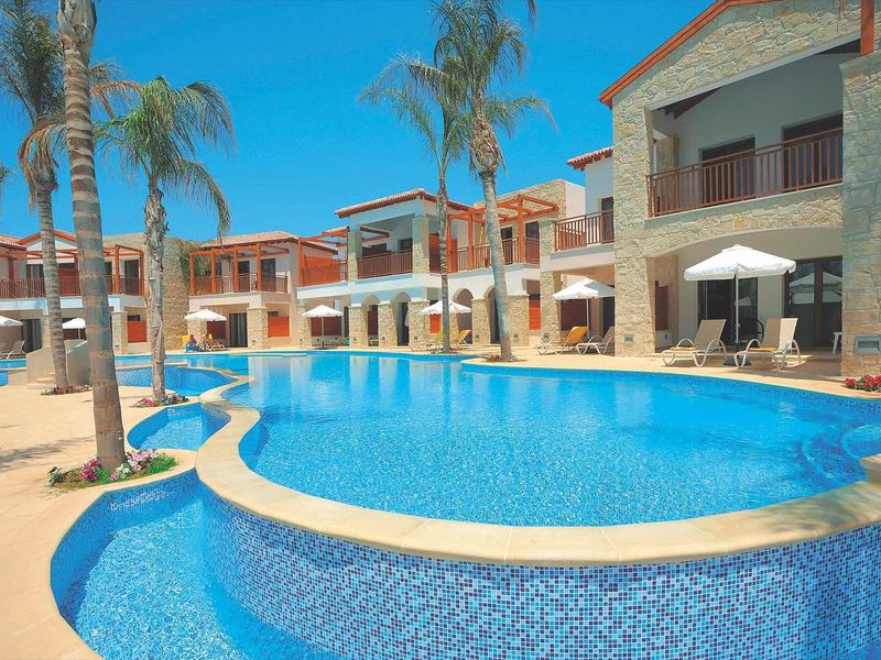 Großer blauer Pool mit palmenumgebenen Steinhäusern unter klarem Himmel.