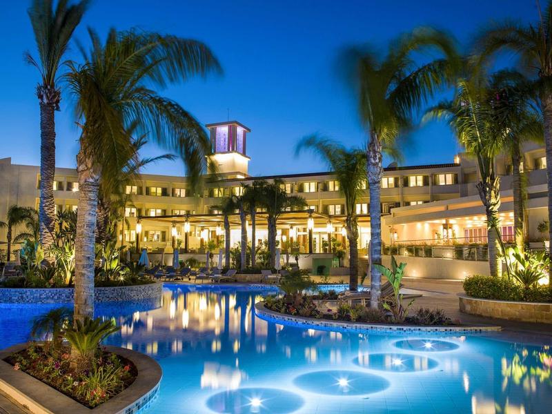 Elegantes Hotel mit beleuchtetem Pool, Palmen und mehrstöckigem Gebäude bei Abenddämmerung.