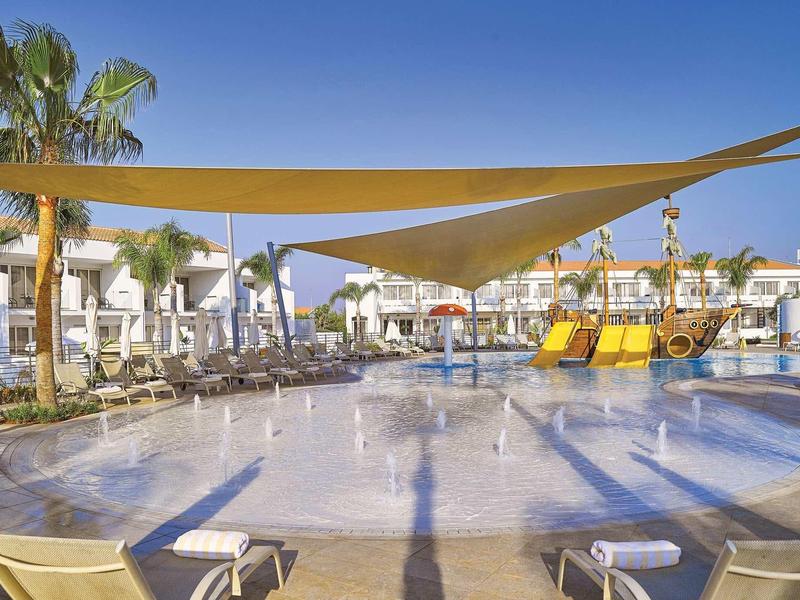 Großes Kinderbecken mit Sonnensegeln, Wasserspielen und gelben Rutschen neben Liegestühlen.