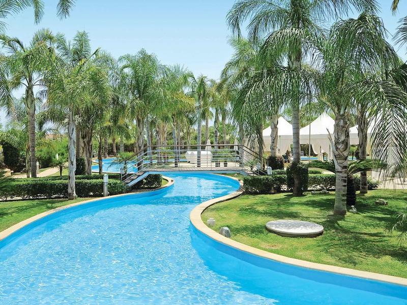 Blauer Pool mit geschwungener Form, umgeben von grünen Palmen und gepflegtem Rasen.