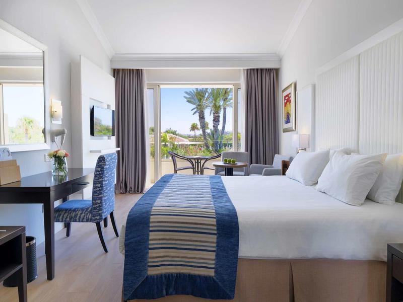 Helles Hotelzimmer mit großem Bett, Schreibtisch, blauer Decke und Blick auf Terrasse mit Palmen.