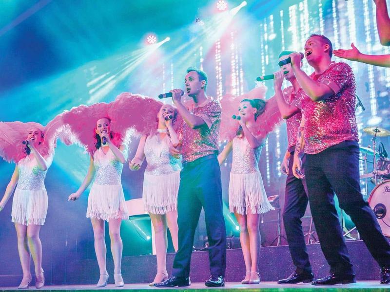 Bunte Bühne mit Tänzerinnen in weißen Kleidern und zwei Männern mit Mikrofonen, bunte Lichtstrahlen.