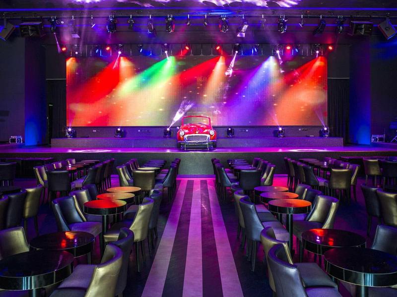 Eleganter Veranstaltungsraum mit bequemen schwarzen Sesseln, runden Tischen und bunter Bühnenbeleuchtung.