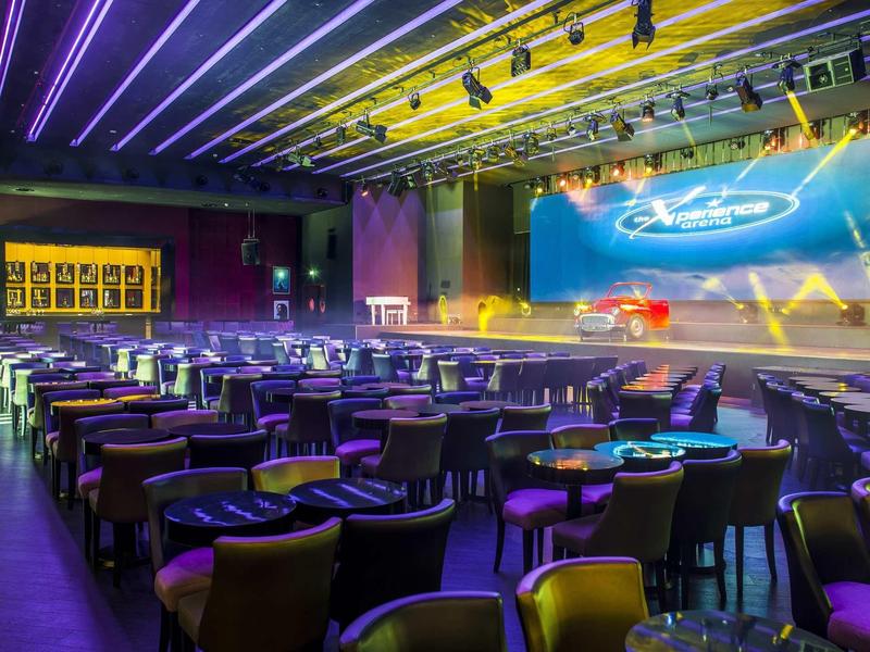 Großer Veranstaltungsraum mit Bühne, mehreren Sitzreihen und bunten Deckenlichtern in Blau und Gelb.