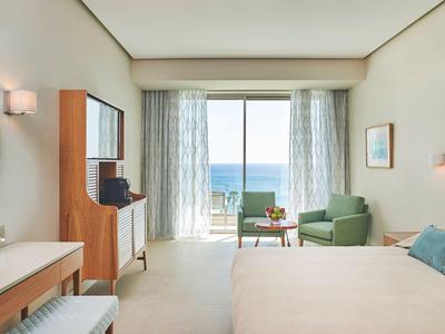 Bright hotel room with double bed, seating area, and sea view through large windows.