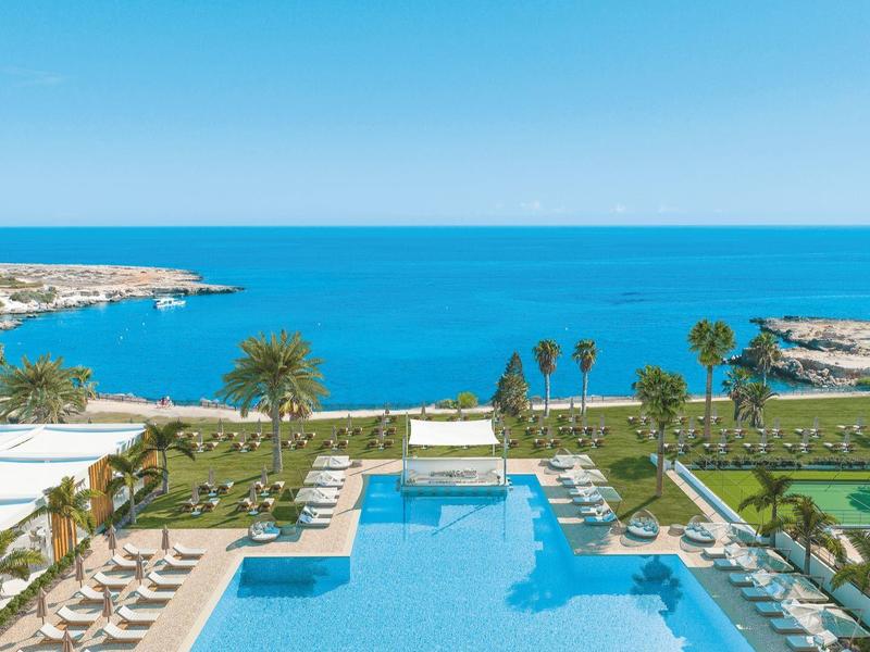 View of a large pool with sun loungers and palm trees with the sea in the background.