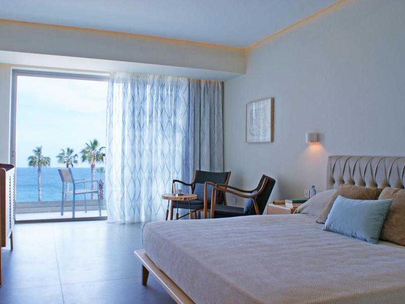 Modern hotel room with sea view, bed, armchairs, and balcony in daylight.