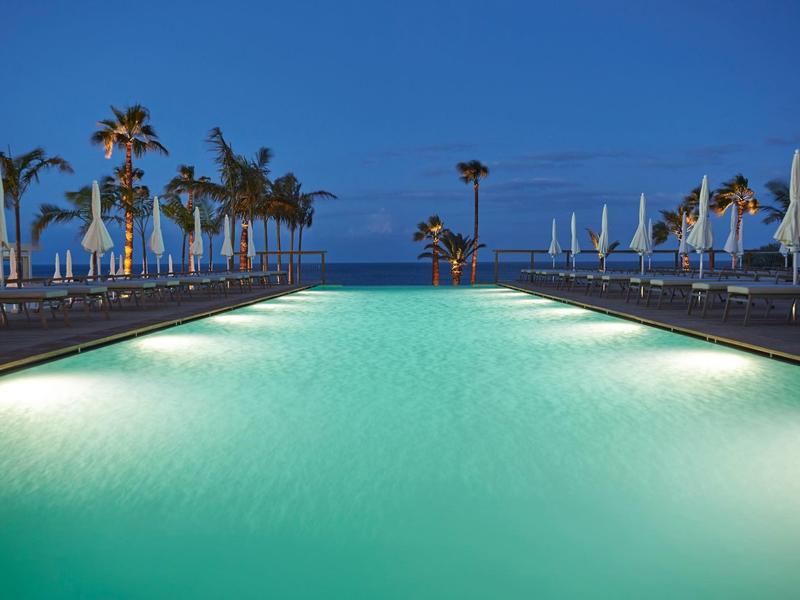 Illuminated pool with loungers and palm trees at dusk in a resort.