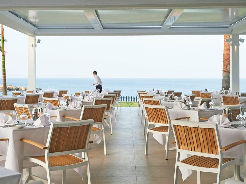 Elegant outdoor restaurant overlooking the sea, with set tables and wooden-metal chairs.