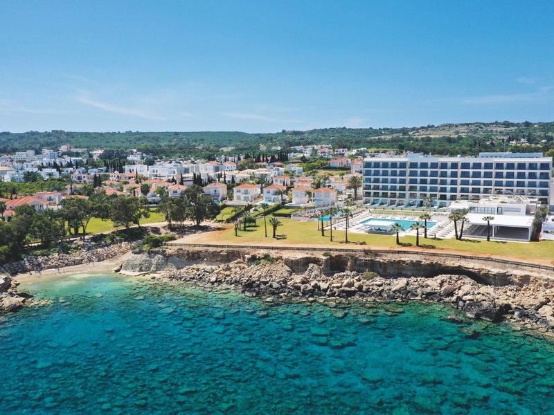 Coastal hotel with pool, surrounded by rocks and clear blue sea under a blue sky.