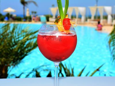 Rotes Cocktailglas mit Fruchtgarnitur vor blauem Pool und sommerlicher Umgebung.