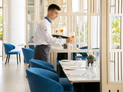 Ein Kellner stellt Gläser auf einen gedeckten Tisch in einem hellen, modernen Restaurant mit blauen Stühlen.