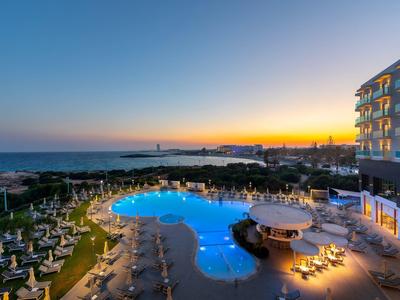 Sonnenuntergang an einem Pool mit blauen Lichtern, Liegestühlen und einem Hotelgebäude am Meer.